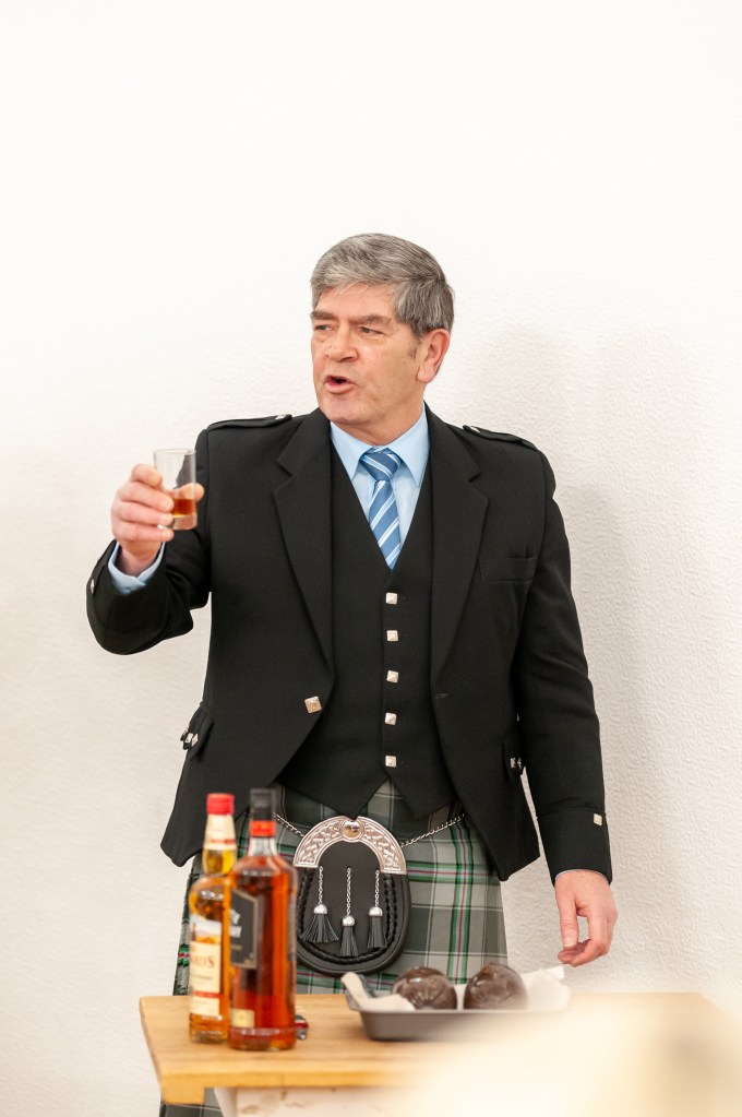A man in a kilt and formal jacket raises a glass of whiskey while giving a speech, with bottles of whiskey and a tray of chocolates on a table.