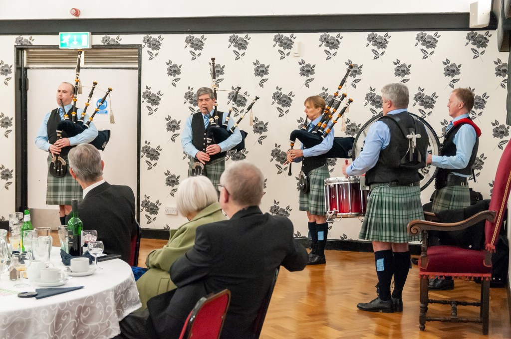 A group of four musicians in traditional Scottish attire, including kilts, performing with bagpipes and a drum in a decorated indoor setting.