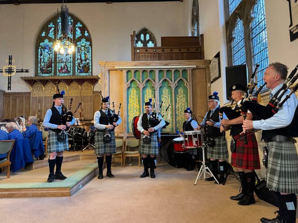 A group of bagpipers wearing traditional Scottish attire, including kilts, perform in a church setting with stained glass windows in the background.